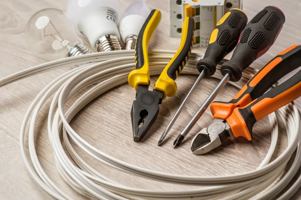 A display of common electrical supplies on a wooden surface, which includes a coil of white wire, various pliers, screwdrivers, LED light bulbs, and a circuit breaker.