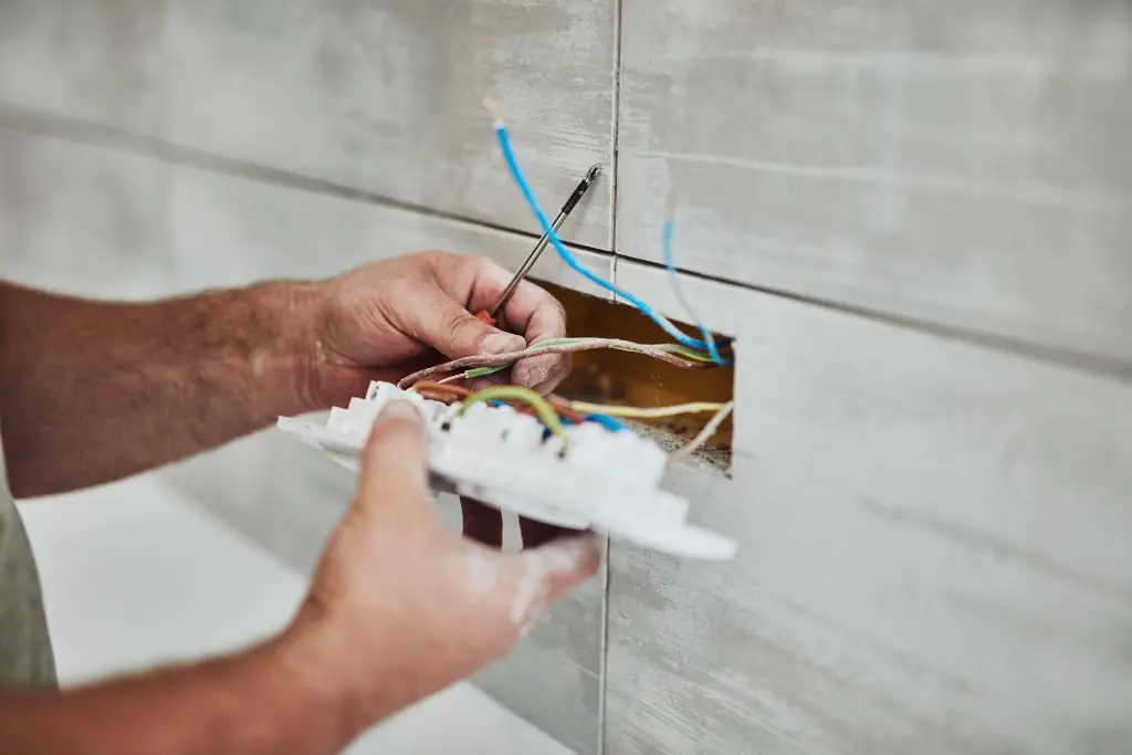 A worker is holding a multi-gang electrical outlet or switch plate, examining the wiring connections before installing it into a tiled wall.